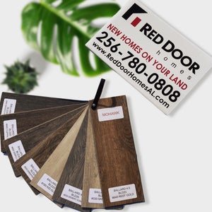 A collection of laminate flooring samples with diverse wood patterns, anchored by a "MOHAWK" tag. In the blurred background, a "RED DOOR HOMES" ad with contact info and a vibrant green plant leaf suggest a lively indoor atmosphere