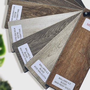 Various brown and beige "MOHAWK" laminate samples displayed, with a "RED DOOR HOMES" business card above them. Details include "NEW HOMES ON YOUR LAND" and contact info. A blurred green plant leaf enhances the indoor setting